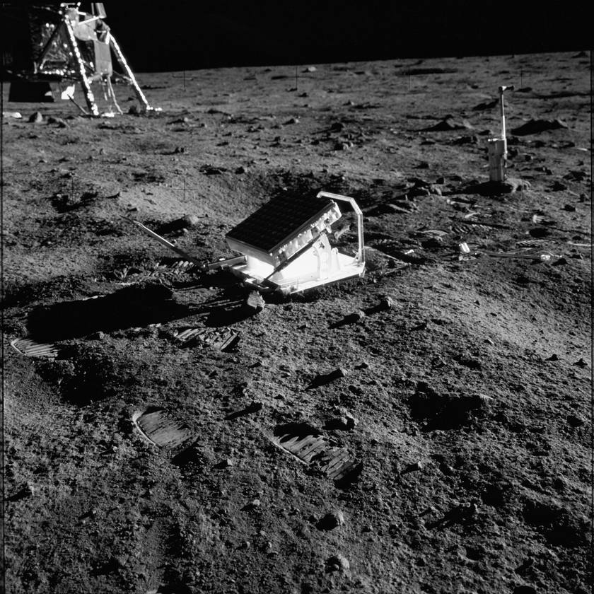 A tilted panel of small square reflectors standing on grey lunar soil, bootprints in the foreground and a lander leg in the corner
